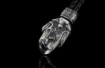 Silver metallic key chain by TOUCHSTONE, featuring a skull and shield design with intricate carvings and a braided black leather strap, reflected on a dark surface.