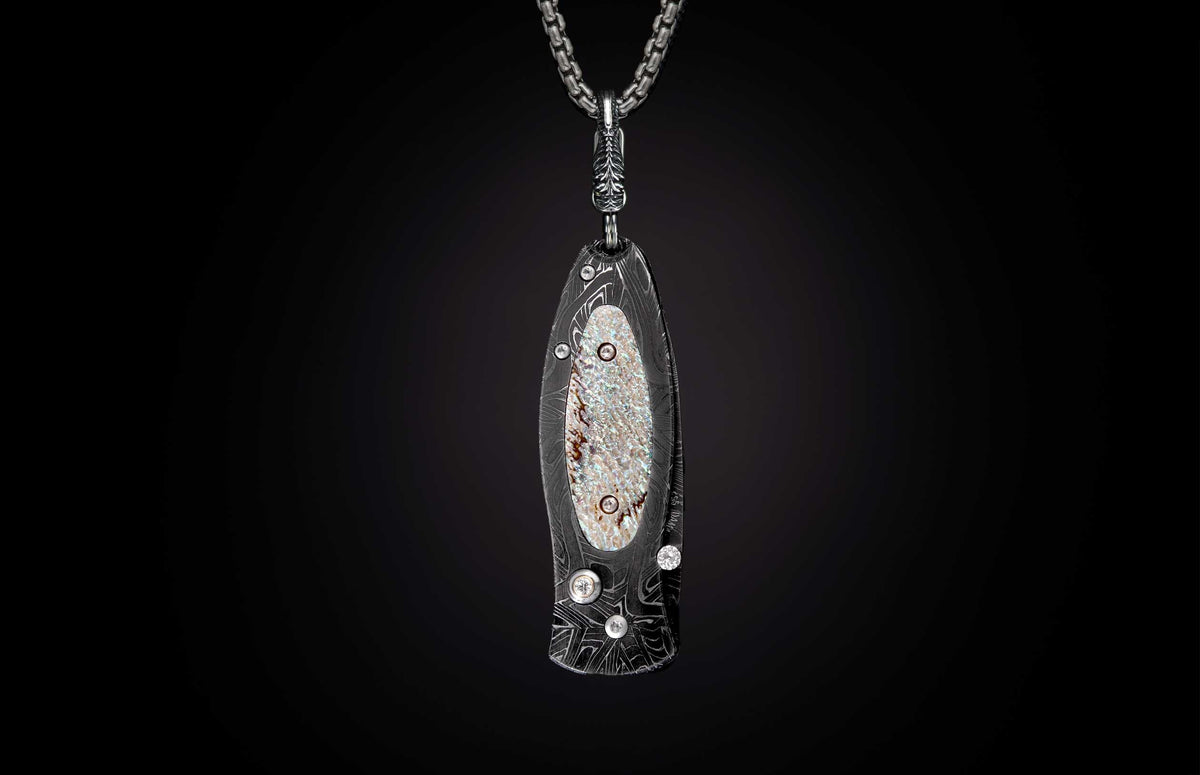 Stylized dark metal knife pendant with wavy Damascus pattern, iridescent mother-of-pearl inlay, and clear stones on a silver chain.