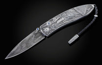 Handcrafted pocket knife from William Henry featuring a stainless steel frame hand-engraved with images of the Statue of Liberty and set with blue sapphires. The blade is hand-forged damascus steel.
