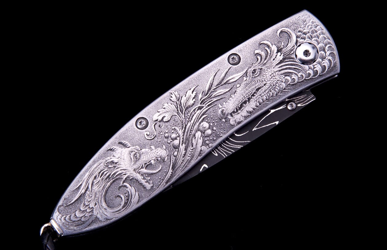 Silver metal folding knife with intricate engraved dragons and foliage on its handle, partially revealing a black blade.