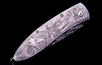 Silver metal folding knife with intricate engraved dragons and foliage on its handle, partially revealing a black blade.