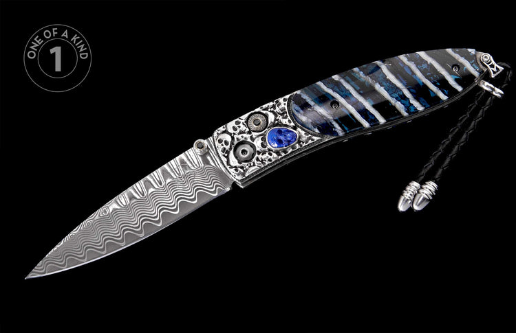 Damascus folding knife with a wavy blade, intricate silver handle featuring skulls, a blue gemstone, blue and white striped inlay, and a black leather lanyard.