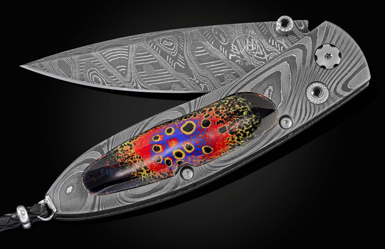 Handcrafted pocket knife from William Henry features a frame in hand-forged Damascus steel, inlaid with enamel artistry by Little Lake Metals. The blade is hand-forged Damascus and the button lock and thumb stud are set with spinel gemstones.