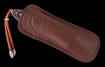 Folding knife with a decorative metal handle, nestled in a brown pebbled leather sheath featuring an embossed logo and braided lanyard.