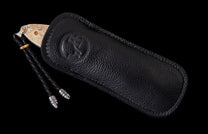 Cream-handled folding knife partially sheathed in a black textured leather case with an embossed logo and braided lanyard.