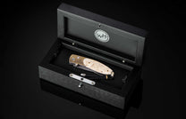 Decorative folding knife with a patterned blade, ornate brass bolster, and light textured handle, in an open black wooden presentation box.