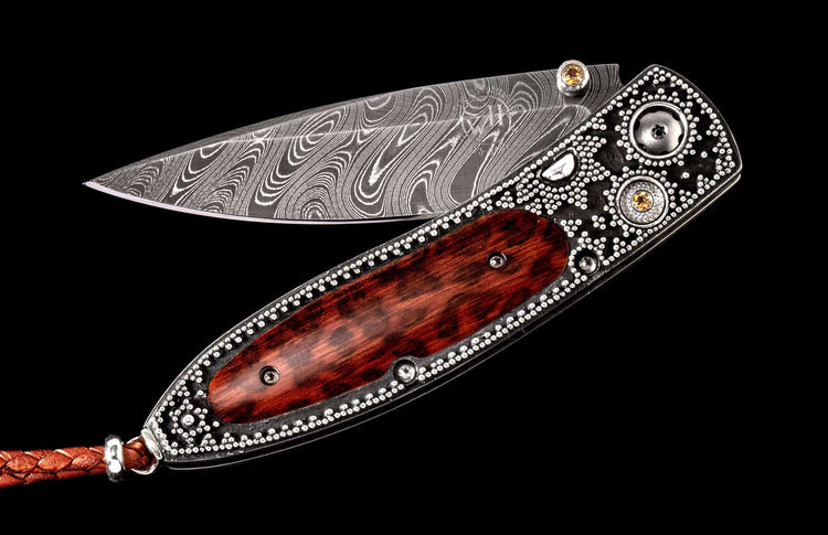 Ornate folding knife with a swirling Damascus steel blade, a dark embellished handle, reddish wood inlay, and yellow gemstones.