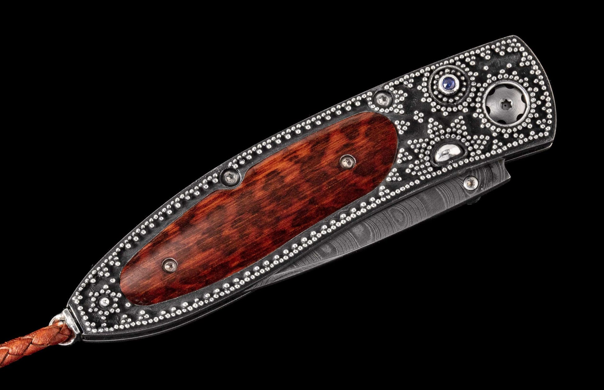 Ornate folding knife with a dark studded handle, reddish-brown wood inlay, blue gem, Damascus steel blade detail, and braided leather lanyard.
