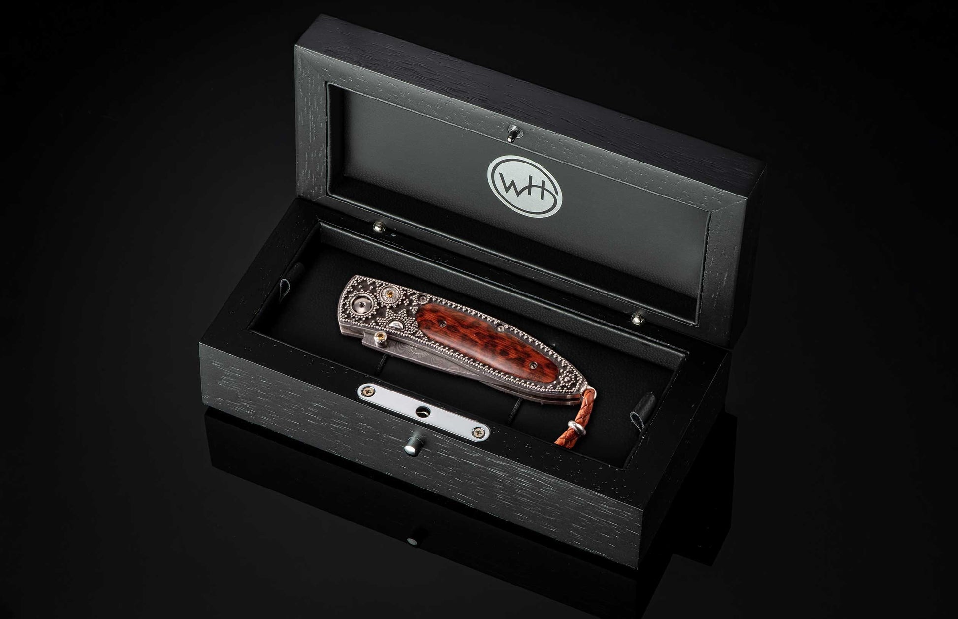 Ornate folding knife with brown wood handle, featuring intricate silver metalwork, displayed in an open black wooden box with a 'WH' logo.