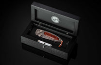 Ornate folding knife with brown wood handle, featuring intricate silver metalwork, displayed in an open black wooden box with a 'WH' logo.