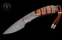 Damascus steel folding knife with swirling patterns, ornate silver and gold engraved handle featuring orange gems, striped brown and white handle scales, and a brown leather lanyard.