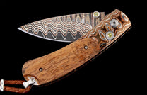 Open folding knife with wavy damascus blade, light brown wood handle, and yellow gemstone accents.