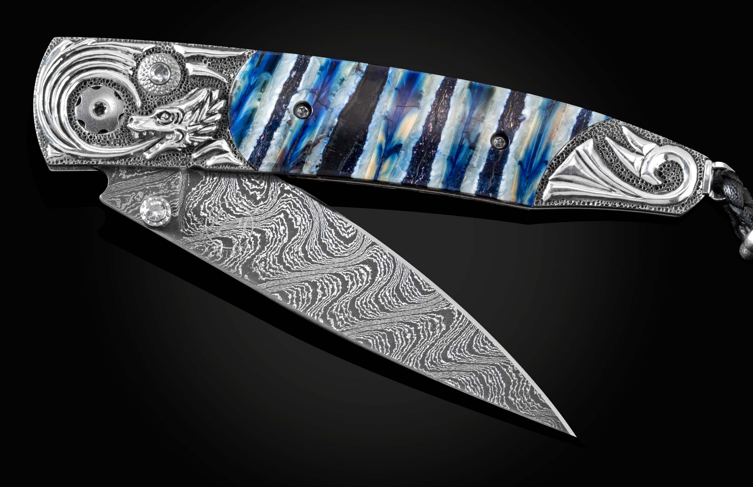 Handcrafted pocket knife from William Henry features a hand-carved sterling silver depicting a dragons head and tail, inlaid with blue Woolly Mammoth tooth, set with white topaz gemstones, and a hand forged Damascus steel blade.