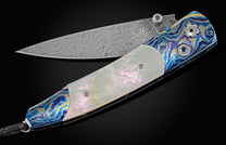 Pocket knife from William Henry featuring a hand forged Damascus steel blade and a frame of hand-forged Ukrainian 'Moku-ti' Damascus, inlaid with Gold Lip Pearl scales. The knife is shown partially open on a black background.