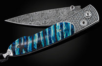 Handcrafted pocket knife from William Henry featuring a laser engraved stainless frame and scales of blue fossil Woolly Mammoth tooth. The blade is hand-forged damascus steel and the button lock and the thumb stud are set with spinel gemstones.