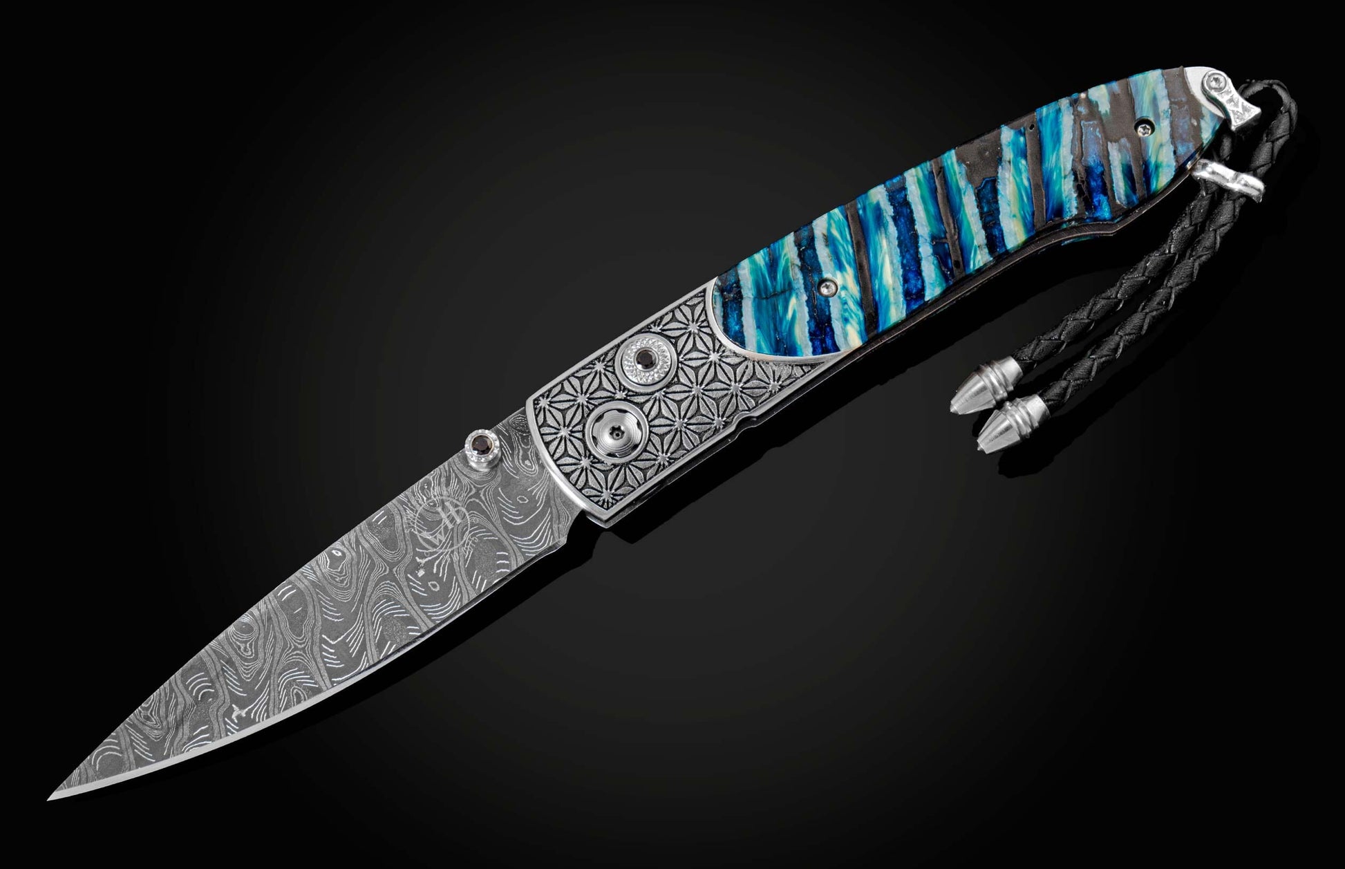 Handcrafted pocket knife from William Henry featuring a laser engraved stainless frame and scales of blue fossil Woolly Mammoth tooth. The blade is hand-forged damascus steel and the button lock and the thumb stud are set with spinel gemstones.