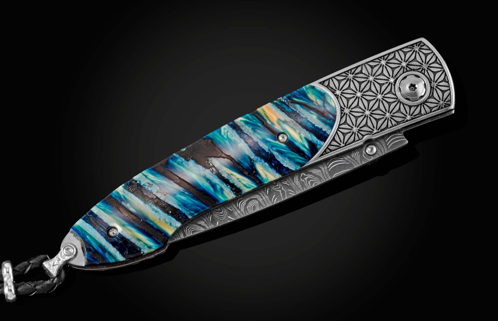 Handcrafted pocket knife from William Henry featuring a laser engraved stainless frame and scales of blue fossil Woolly Mammoth tooth. The blade is hand-forged damascus steel and the button lock and the thumb stud are set with spinel gemstones.