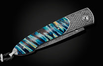 Handcrafted pocket knife from William Henry featuring a laser engraved stainless frame and scales of blue fossil Woolly Mammoth tooth. The blade is hand-forged damascus steel and the button lock and the thumb stud are set with spinel gemstones.