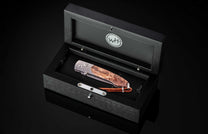 Folding knife with a silver frame and burled wood handle, displayed in an open black wooden presentation box with a 'WH' logo and a brown braided leather lanyard.