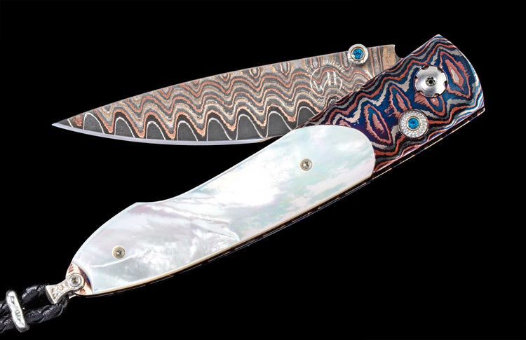 Knife with a wavy Damascus blade, blue and orange patterned handle, iridescent mother-of-pearl, blue gemstones, and a black braided cord.
