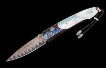 Folding knife with a wavy patterned blade, iridescent mother-of-pearl, colorful swirling handle, blue gemstones, and a braided leather lanyard.