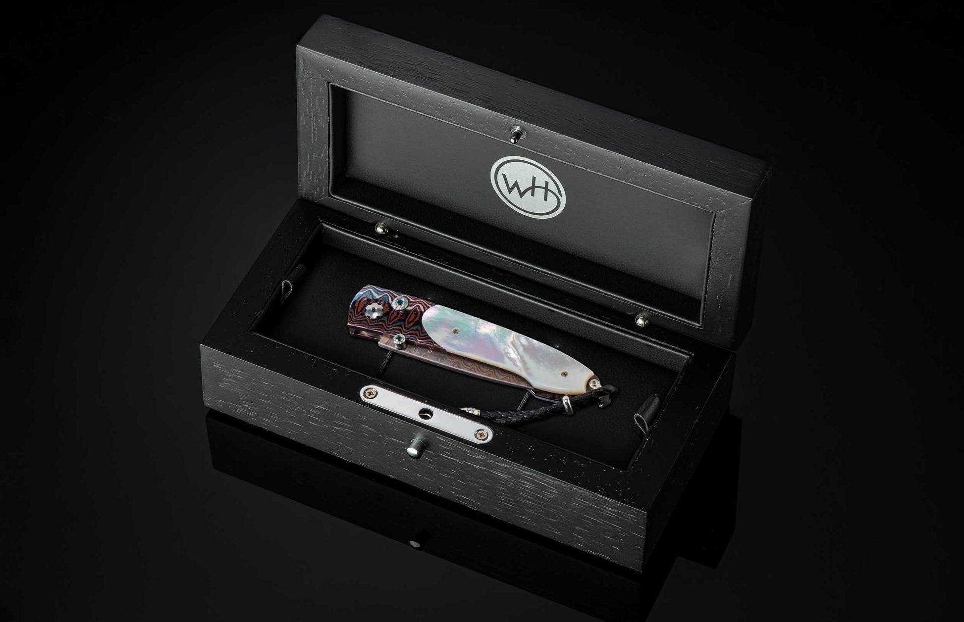 Ornate folding knife with iridescent mother-of-pearl and patterned steel handles, accented with blue gems, displayed in an open black wooden box with a 'WH' logo.