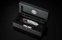 Ornate folding knife with iridescent mother-of-pearl and patterned steel handles, accented with blue gems, displayed in an open black wooden box with a 'WH' logo.
