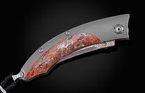 Handcrafted pocket knife from William Henry features a titanium frame inlaid with orange and red zinc matrix Apple Coral. The button lock and the thumb stud have been set with citrine gemstones and the hand forged Damascus steel blade has a copper wave pa