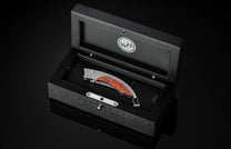 Handcrafted pocket knife from William Henry features a titanium frame inlaid with orange and red zinc matrix Apple Coral. The button lock and the thumb stud have been set with citrine gemstones and the hand forged Damascus steel blade has a copper wave pa