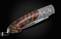 Candcrafted pocket knife from William Henry featuring hand-engraved grouse and quail with copper and 24K gold inlaid with fossil Woolly Mammoth tusk and set with spinel gem stones.. The blade is hand-forged damascus steel.