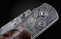 Candcrafted pocket knife from William Henry featuring hand-engraved grouse and quail with copper and 24K gold inlaid with fossil Woolly Mammoth tusk and set with spinel gem stones.. The blade is hand-forged damascus steel.