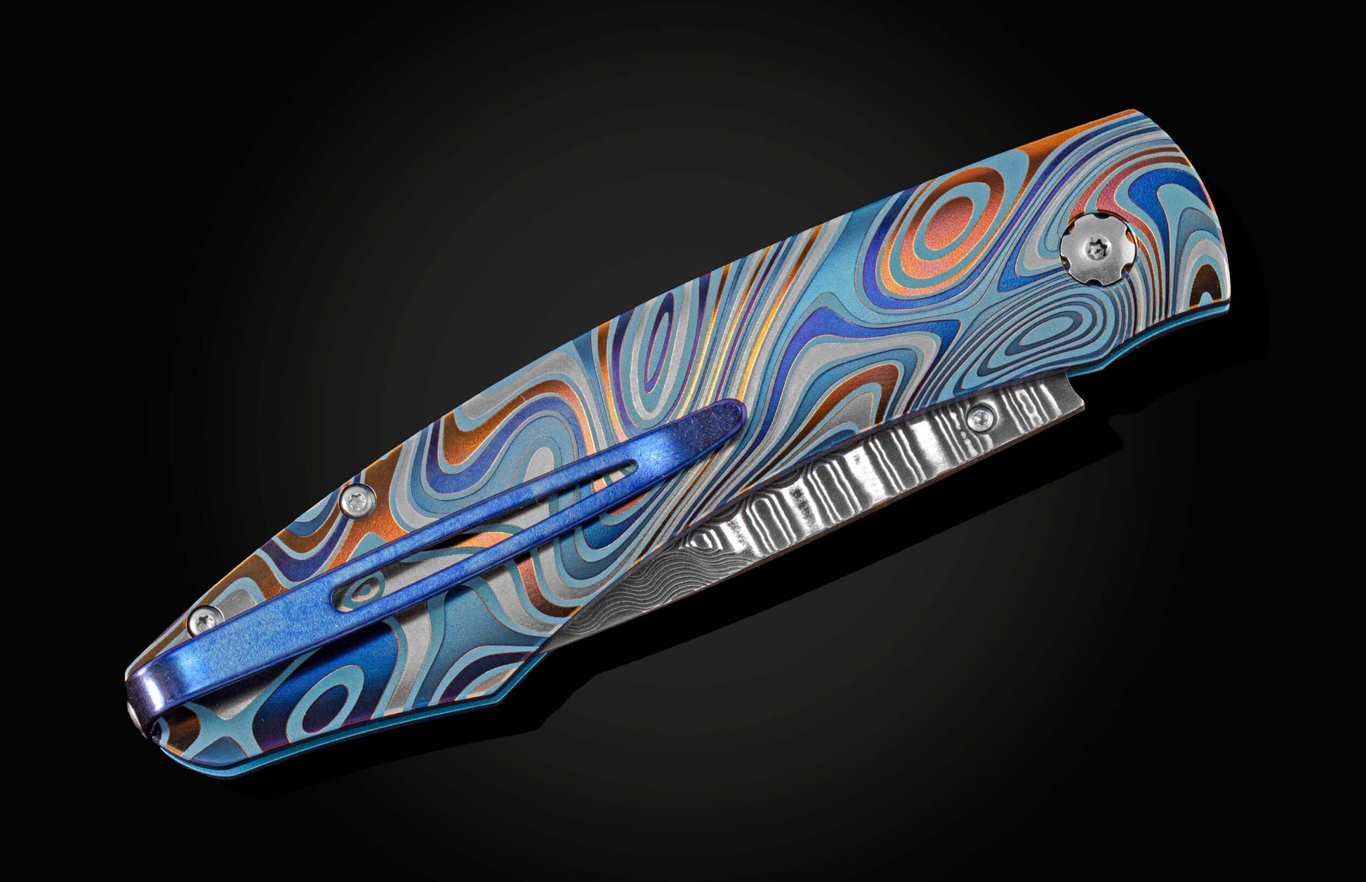 Unique pocket knife from William Henry in swirling colors on laser-engraved titanium with hand-forged damascus blade and blue topaz gemstones. The knife is displayed closed on a black background.