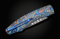 Unique pocket knife from William Henry in swirling colors on laser-engraved titanium with hand-forged damascus blade and blue topaz gemstones. The knife is displayed closed on a black background.