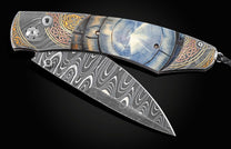 Handcrafted pocket knife from William Henry features a hand-forged damascus steel frame with 24K and 22K yellow gold, 18K green gold, and copper with spinel gemstones., inlaid with fossil Woolly Mammoth tusk. The blade is hand forged damascus steel which 