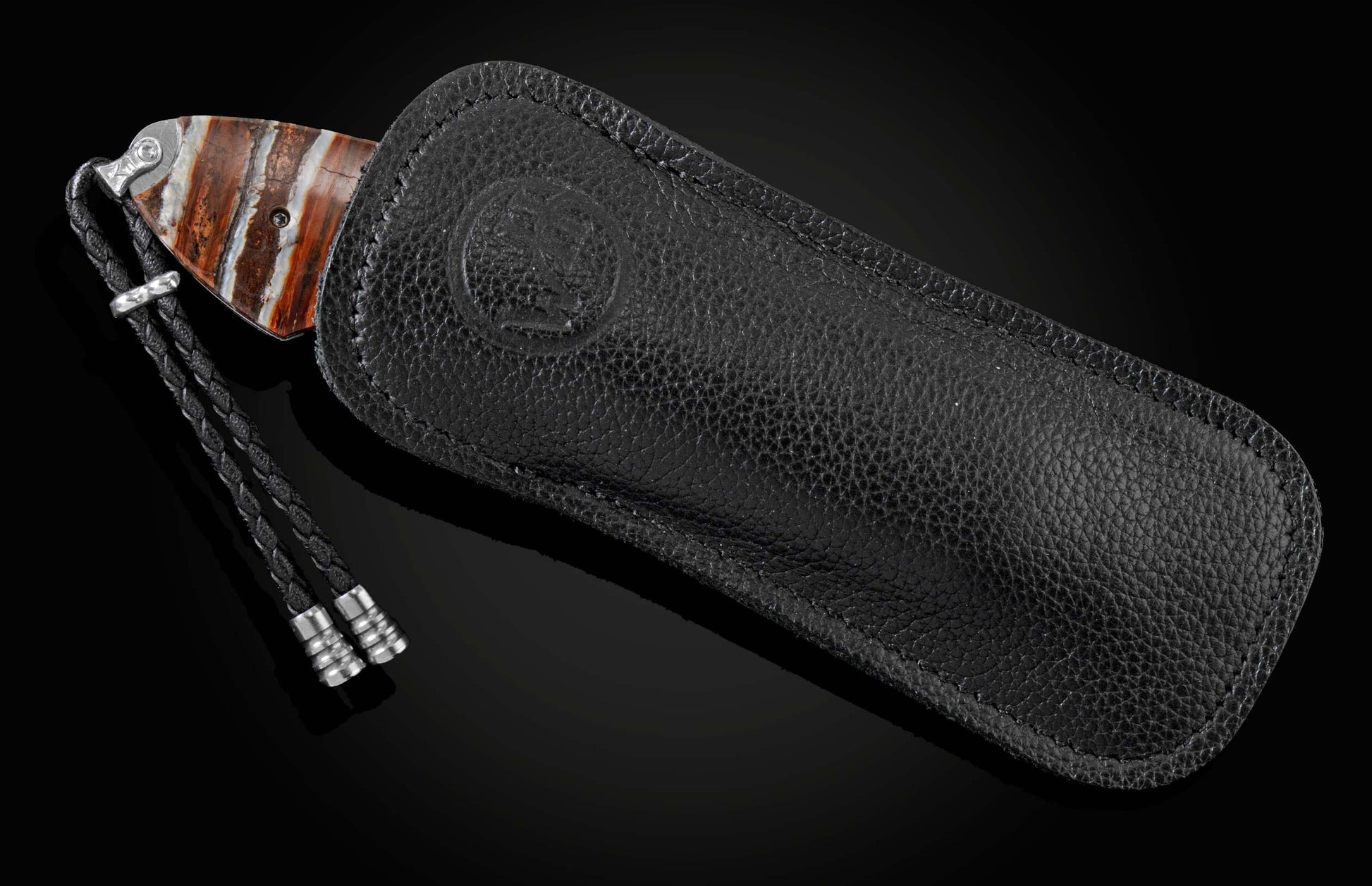 Knife with a striped brown, white, and orange handle, silver bolster, partially in a black pebbled leather sheath with embossed logo, and a black braided lanyard.