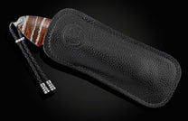 Knife with a striped brown, white, and orange handle, silver bolster, partially in a black pebbled leather sheath with embossed logo, and a black braided lanyard.