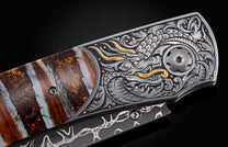 Ornate folding knife with a silver and gold dragon engraving, featuring a striped natural stone handle and a dark patterned blade, side view.
