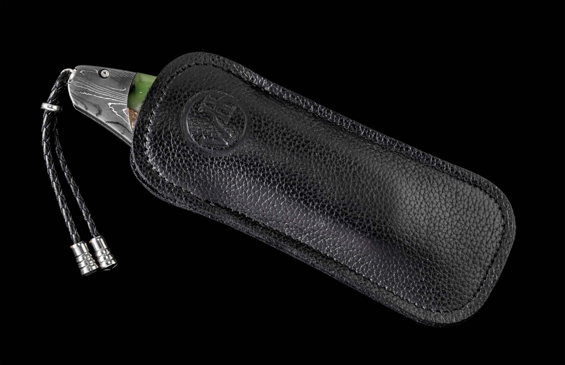 B12 Eagle II knife with patterned blade and green accent, partially in a black pebbled leather sheath with an embossed logo and braided lanyard with silver tips.