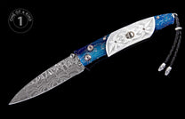 A decorative folding knife with a Damascus steel blade, a blue handle with gold marbling, and a white carved handle featuring a silver skull and a black leather lanyard.