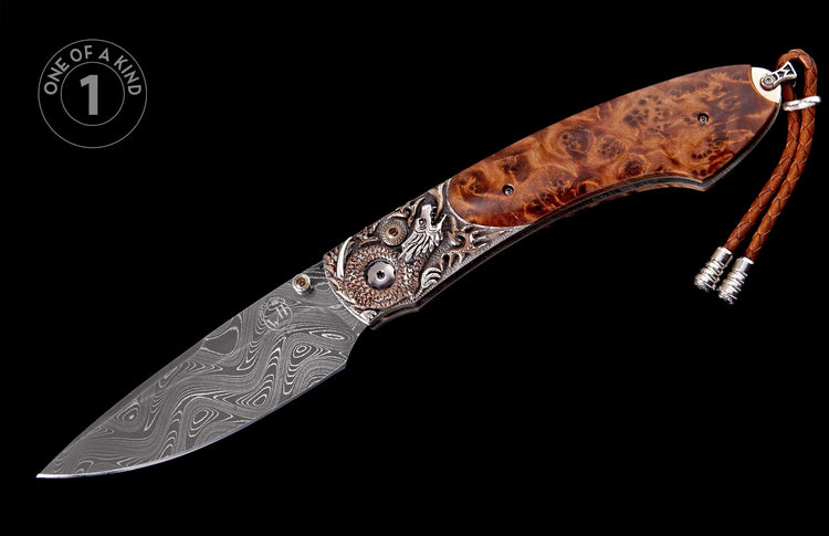 Damascus folding knife with a burled wood handle, intricate dragon motif, brass accents, and a braided leather lanyard, labeled 'ONE OF A KIND'.