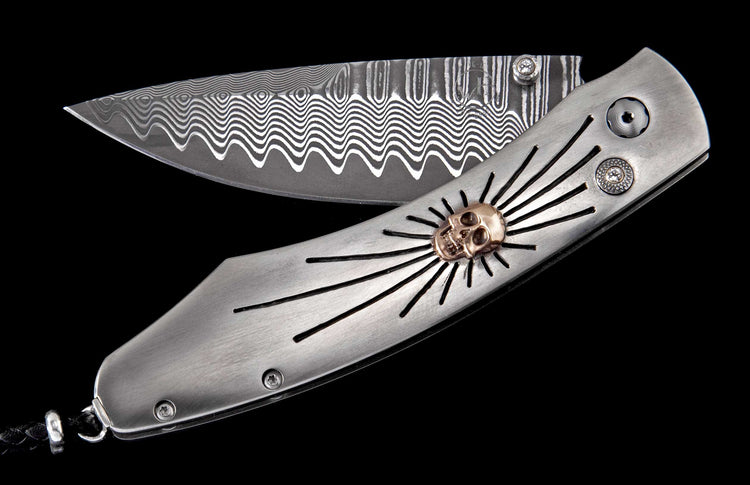 Silver metal folding knife with a wavy Damascus steel blade, a bronze skull emblem with radiating lines, and two sparkling gems on the handle.