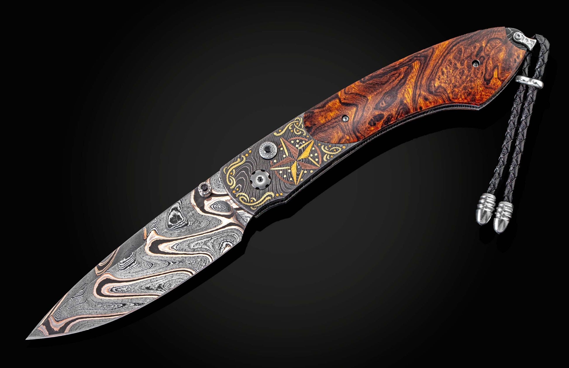 Custom folding knife with a damascus steel blade and a patterned wooden handle, featuring ornate gold and bronze star-shaped engraving and a braided leather lanyard.