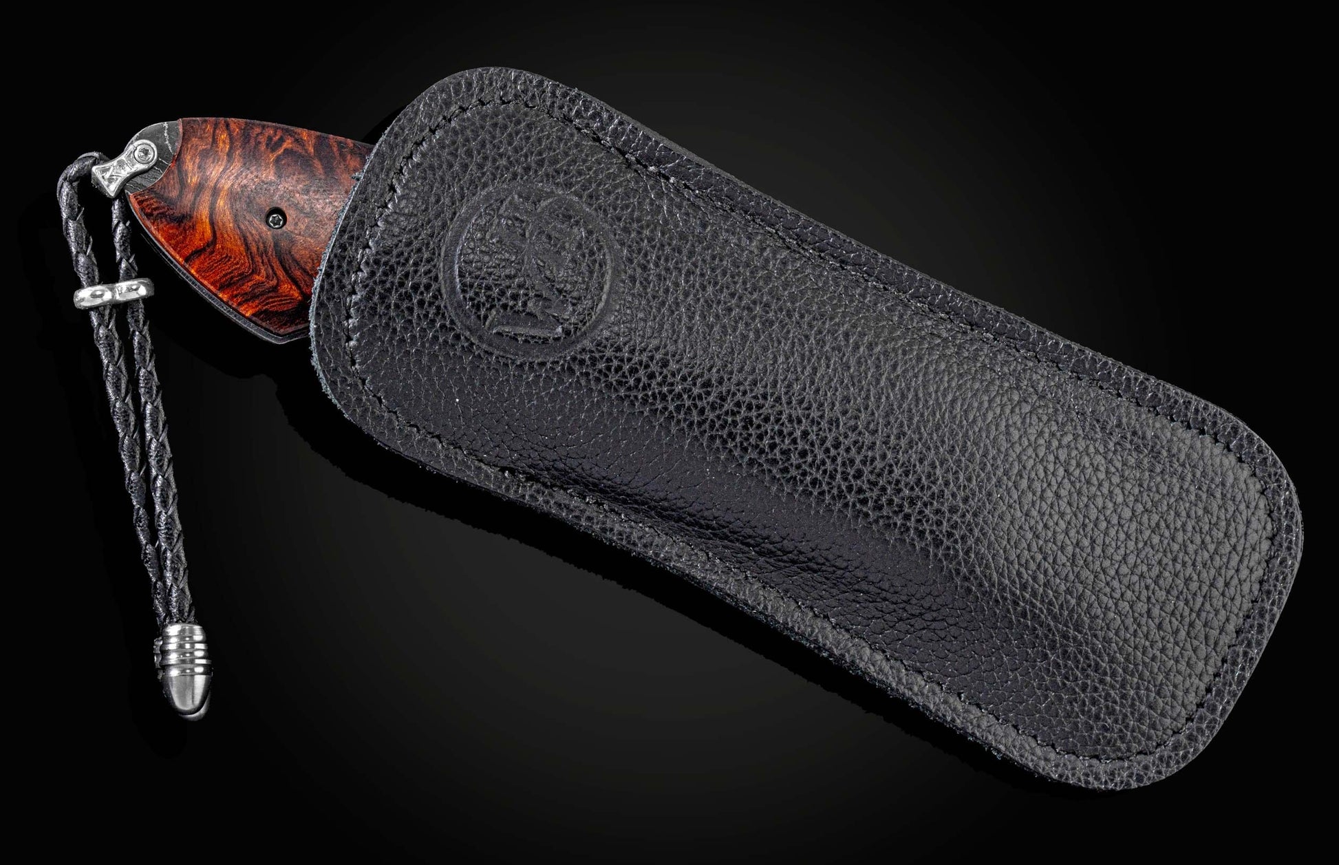 Knife with a reddish-brown burl wood handle and braided black leather lanyard, partially sheathed in black pebbled leather with an embossed logo.