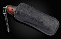 Knife with a reddish-brown burl wood handle and braided black leather lanyard, partially sheathed in black pebbled leather with an embossed logo.