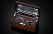 A folding knife with a richly grained brown wooden handle, gold star, and ornate accents, with a Damascus steel blade and black lanyard, displayed in an open dark wood box.