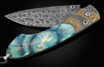 Hand crafted pocket knife from William Henry with a hand forged Damascus steel blade features hand-engraved bolsters with 22K and 24K yellow gold, 18K green gold, and copper inlays, inlaid with a brilliant blue ring-cut of 10,000 year-old fossil Woolly Ma