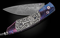 Ornate Damascus steel knife by SEWE featuring a purple and blue iridescent handle with intricate silver engraving, gemstone accents, and a braided leather lanyard, shown open.