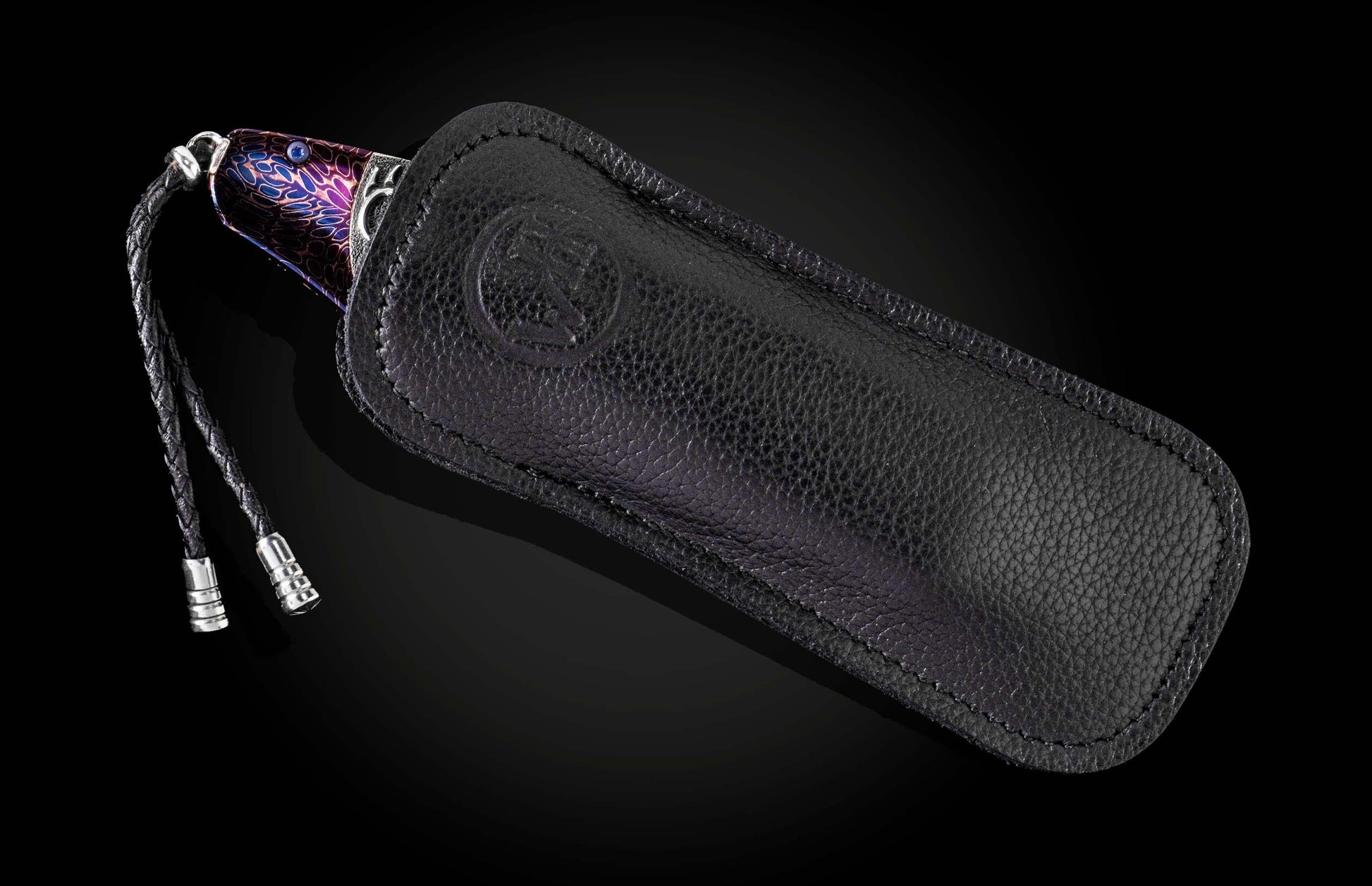 Colorful patterned knife by SEWE with a blue gem, partially sheathed in a black textured leather case with an embossed logo and braided lanyard.