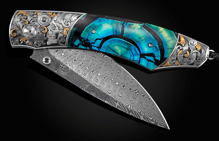 Handcrafted pocket knife from William Henry features hand-engraved skulls and decorative ornamentation with 24K gold and silver inlays and blue woolly mammoth tusk scales, a hand-forged Turkish Damascus steel blade, and spinel gemstones.