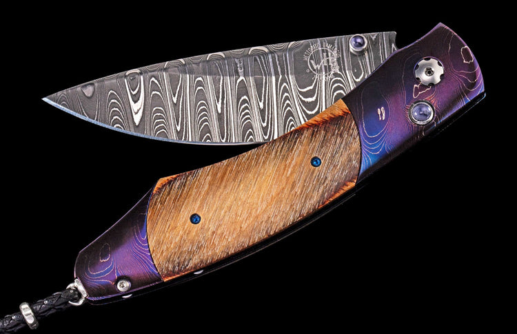 Partially open folding knife with a swirled Damascus blade, iridescent purple and textured wood handle, blue studs, and a braided black leather lanyard.